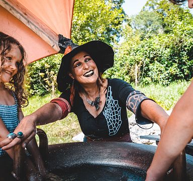 Eine Frau mit Hexenhut lächelt neben einem jungen Mädchen im Freien. Sie sind an einem großen Metallkessel, umgeben von grünen Bäumen.