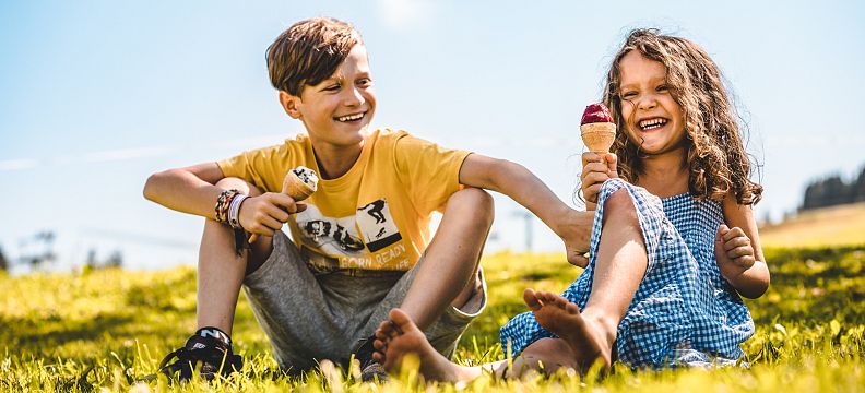 Zwei fröhliche Kinder sitzen auf einer Wiese und genießen Eiscreme an einem sonnigen Tag. Das Mädchen trägt ein blaues Kleid, der Junge ein gelbes T-Shirt.