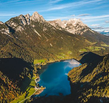 Luftaufnahme eines malerischen Sees in den Alpen, umgeben von bewaldeten Hügeln und majestätischen Bergen unter blauem Himmel bei Tageslicht.