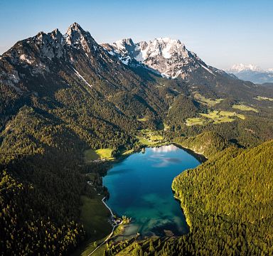 Luftaufnahme eines smaragdgrünen Sees umgeben von dichten Wäldern und beeindruckenden Bergen unter blauem Himmel in einer alpinen Landschaft.