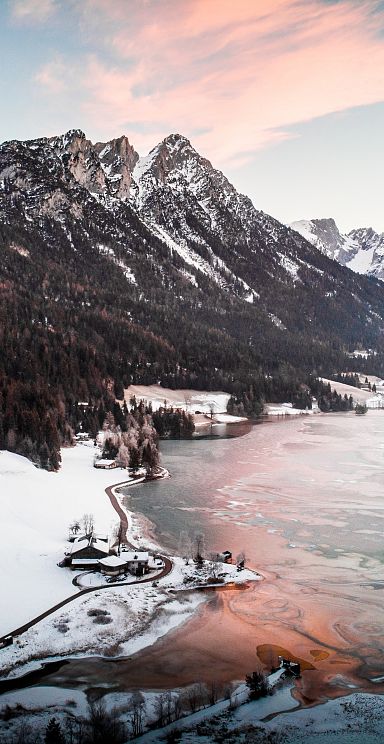 A snow-covered landscape with the Wilder Kaiser mountains in the background, overlooking a partially frozen lake and surrounded by forests at sunset.