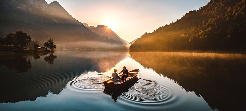 Two people rowing a boat on a calm lake at sunrise, surrounded by mountains, with the sun rising between peaks creating a serene and reflective atmosphere.