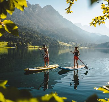 Zwei Personen stehen auf Paddleboards auf einem ruhigen See, umgeben von grünen Wäldern und Bergen im Hintergrund, bei sonnigem Wetter.