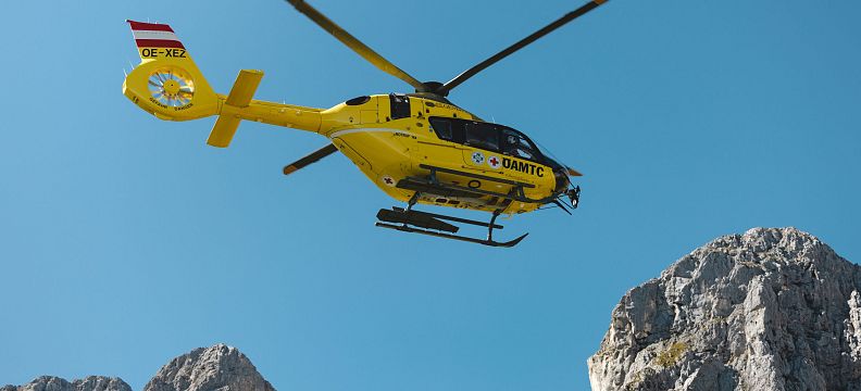 A yellow rescue helicopter soars above the rocky peaks of the Wilder Kaiser against a backdrop of clear blue sky, showcasing dynamic alpine rescue in action.