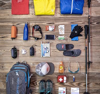 Flach gelegte Wanderutensilien auf Holz: Wasserflaschen, Handschuhe, Stirnlampe, Snacks, Geld, Karte, Handy, Stöcke, Rucksack, Kappe und Erste-Hilfe-Set.