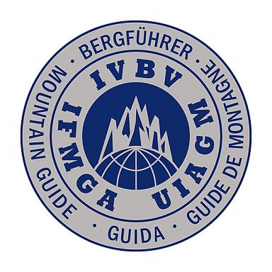 Logo der internationalen Bergführerverband mit Bergspitzen und Schriftzug in verschiedenen Sprachen auf rundem Abzeichen.