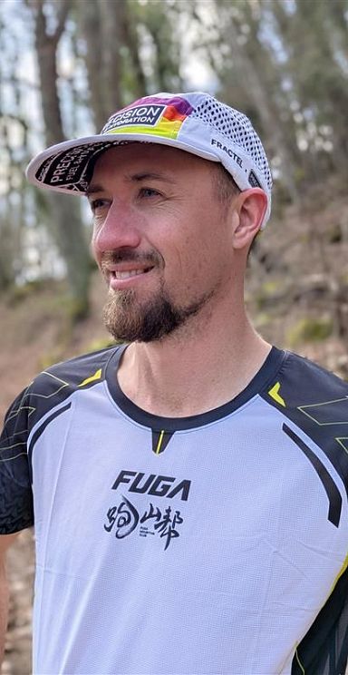 Ein lächelnder Mann im Wald trägt ein Sportshirt und eine Schirmmütze. Der Hintergrund zeigt unscharfe Bäume und einen erdigen Weg.