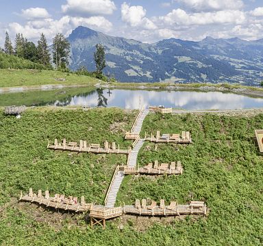 A scenic area in Wilder Kaiser, Tirol, with a tranquil pond, surrounding greenery, and mountains in the background on a sunny day.