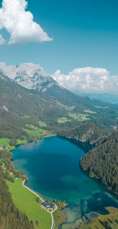 Panorama eines Bergsees umgeben von grünen Wäldern und majestätischen Bergen unter blauem Himmel.
