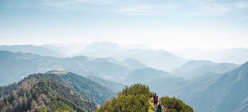 Zwei Wanderer auf einem schmalen Pfad über grüne Bergkämme, umgeben von majestätischen bergigen Landschaften unter einem klaren blauen Himmel.