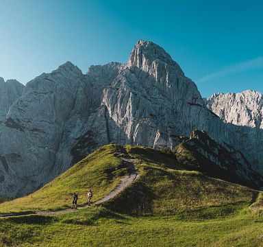 Wanderer auf einem grasbewachsenen Pfad vor einer beeindruckenden Felsformation unter klarem blauem Himmel; typisches alpines Panorama.