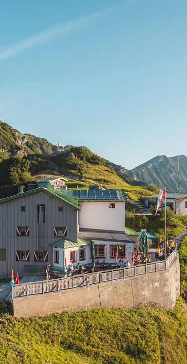 Bergsteigerhütte auf einem grünen Berghang mit Blick auf umliegende Gipfel bei klarem Himmel. Die Hütte ist von einem Pfad erreichbar.