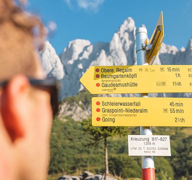 Ein Wanderer schaut auf Wegweiser im Vordergrund, die verschiedene Ziele in den Alpen anzeigen. Im Hintergrund erheben sich beeindruckende Berggipfel.