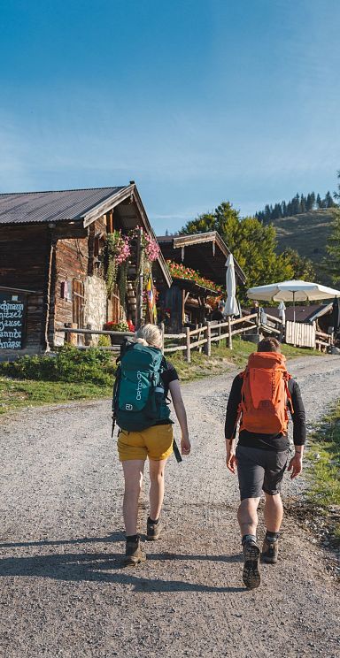 Two hikers with backpacks walk on a gravel path towards a mountain hut in the Wilder Kaiser, surrounded by green meadows and blue sky, capturing a peaceful outdoor scene.