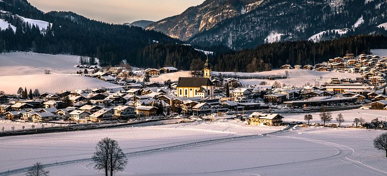 Winterliches Bergdorf in Tirol umgeben von schneebedeckten Hügeln und Wäldern, eine Kirche steht zentral, unter der Abenddämmerung, idyllisch gelegen.