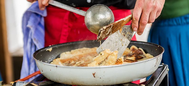 Ein Koch bereitet in einer Pfanne auf einem Herd ein Gericht zu, vermutlich Crêpes oder Pfannkuchen. Die Szene findet auf einem Markt oder Fest statt.