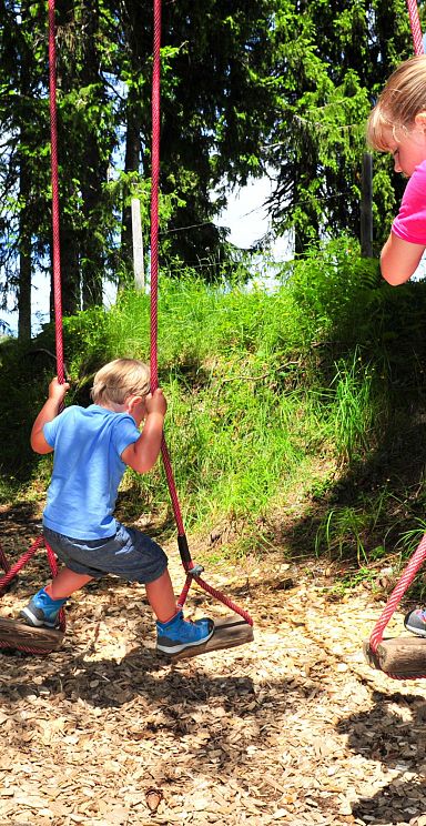 Two children enjoy swings tied to trees in a forest at Wilder Kaiser. The sun filters through the trees, creating a lively and adventurous atmosphere.