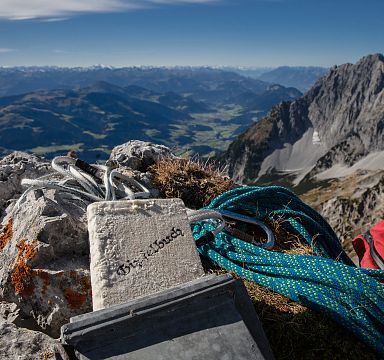 Summit view with climbing gear on rocky peak, overlooking expansive alpine landscape and distant mountain ranges under a clear blue sky.