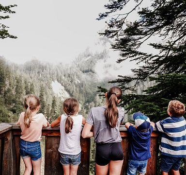 Fünf Kinder stehen auf einem Holzsteg und blicken auf einen bewaldeten, nebelverhangenen Hügel in einem Naturgebiet. Die Szene wirkt ruhig und idyllisch.