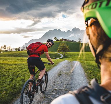 Two mountain bikers on a scenic trail, one in focus wearing a helmet, with the Wilder Kaiser mountains majestically rising in the background under a cloudy sky.