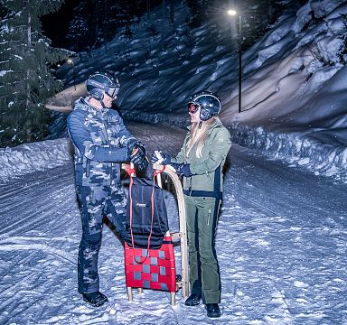 Two people in winter gear stand with a sled on a snowy, illuminated path lined with snow-covered trees.