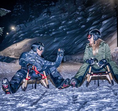 Two people in winter gear sit on sleds, smiling and enjoying a night sledding adventure on a snow-covered path surrounded by trees at Wilder Kaiser.