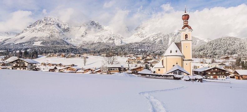 Snow-covered landscape with a church and the Wilder Kaiser mountains in the background, under a partly cloudy sky.