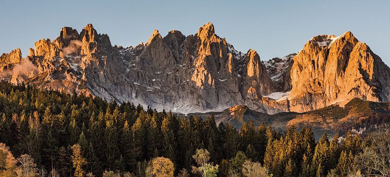 Sunlit mountain peaks rise above lush green forests and fields, with a clear blue sky enhancing the majestic landscape.