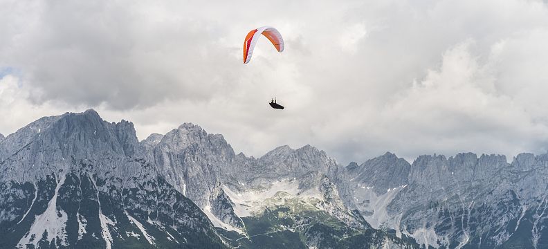 Ein Gleitschirmflieger schwebt über einer alpinen Berglandschaft mit schneebedeckten Gipfeln und bewaldeten Tälern unter einem bewölkten Himmel.