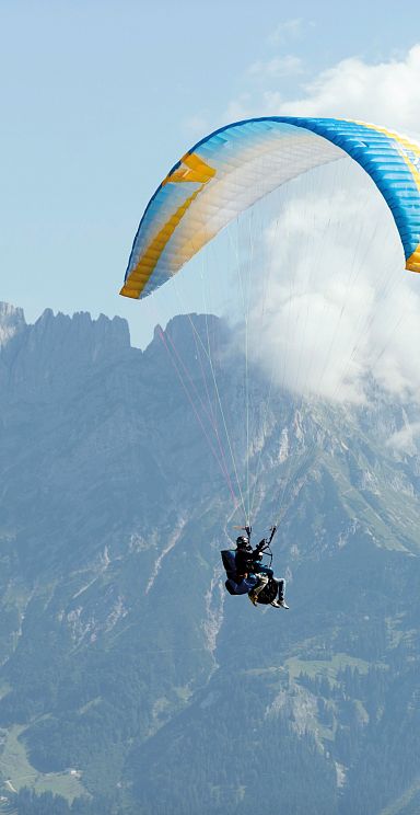 Ein Gleitschirmflieger schwebt über einer alpinen Landschaft mit beeindruckenden Berggipfeln im Hintergrund. Der Himmel ist teils bewölkt.