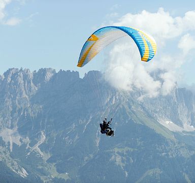 Ein Gleitschirmflieger schwebt über einer alpinen Landschaft mit beeindruckenden Berggipfeln im Hintergrund. Der Himmel ist teils bewölkt.