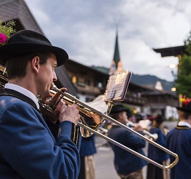 Traditional Austrian brass band performing in a village near the Wilder Kaiser, featuring musicians in blue attire and traditional hats, with a picturesque church steeple in the background.