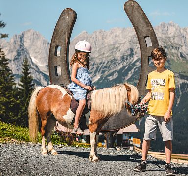 Children pose with a pony against the majestic backdrop of the Wilder Kaiser mountains on a sunny day, highlighting outdoor fun and stunning alpine scenery.