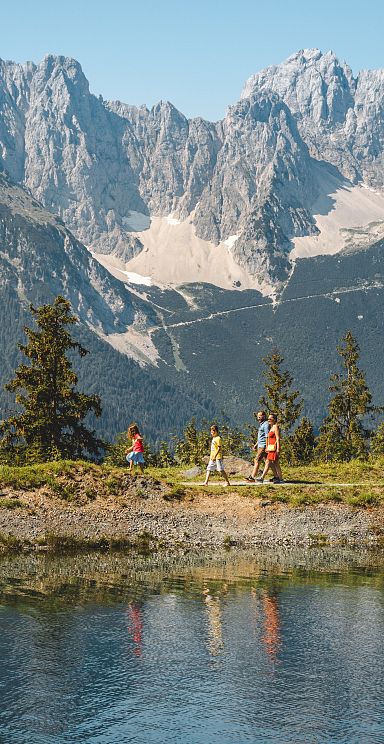 People hike along a scenic mountain path by a lake, with majestic peaks of the Wilder Kaiser range rising in the background under a clear blue sky.