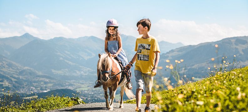 Two children with a pony on a mountain path, surrounded by lush greenery and clear skies, with the majestic Wilder Kaiser mountain range in the distance.