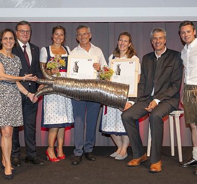 Group of people in formal and traditional attire on stage, holding awards and a large, decorative item, smiling for a photo.