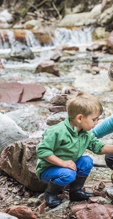 A woman and a child are stacking stones by a stream in the Wilder Kaiser region, surrounded by rocks and greenery. The atmosphere is peaceful and natural.