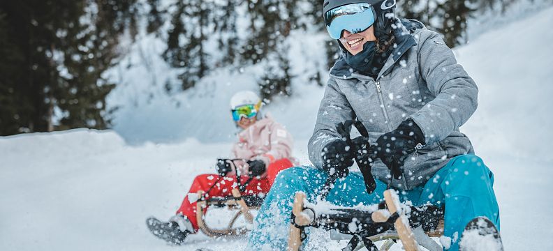 Two people wearing helmets and winter gear are sledding joyfully down a snowy slope surrounded by trees.