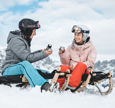 Zwei Personen sitzen lächelnd auf Schlitten im Schnee und genießen warme Getränke, umgeben von einer winterlichen Berglandschaft unter blauem Himmel.