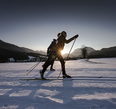 Zwei Personen beim Skilanglauf im Schnee, umgeben von Bergen und einem klaren Himmel, während die Sonne tief am Horizont steht und Lichtstrahlen erzeugt.