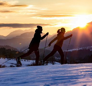 Silhouetted skiers move across snow at sunrise in the Wilder Kaiser, with mountains in the background and a golden sun illuminating the scene.