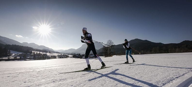 Two skiers cross-country skiing on a sunny day at Wilder Kaiser with mountains and clear blue sky.