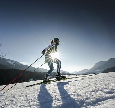 A skier descends a snowy slope under a clear sky, with the sun shining brightly behind, creating a starburst effect. The Wilder Kaiser mountain range is in view.