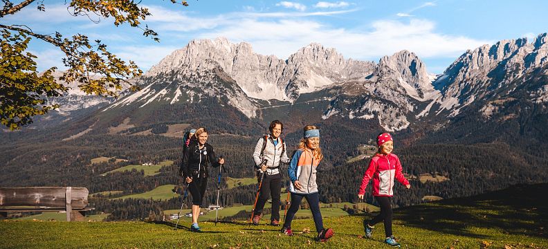 Hikers walking on a green trail with the Wilder Kaiser mountains in the background under a clear blue sky.