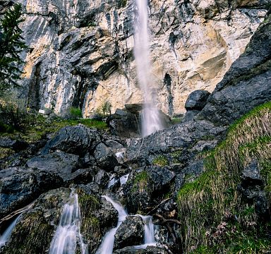Waterfall cascading down rocky cliffs in a lush, green landscape, surrounded by boulders and vegetation, under a clear, blue sky.