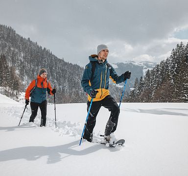 Two people snowshoeing through deep snow in a scenic, snow-covered landscape with trees and mountains in the background at Wilder Kaiser.