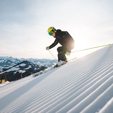 Skifahrer in schwarzer Kleidung fährt bei Sonnenschein eine präparierte Piste hinunter, im Hintergrund verschneite Berge unter klarem Himmel.