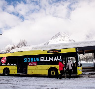 Ein gelber Skibus mit der Aufschrift "SKIBUS ELLMAU" steht an einer Bushaltestelle im Schnee. Eine Person steigt ein, umgeben von schneebedeckter Landschaft.