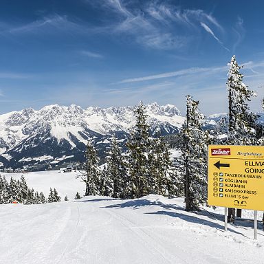 Snow-covered Wilder Kaiser mountain range with a signpost showing directions to Ellmau and Going, set against a clear blue sky and snow-laden trees.
