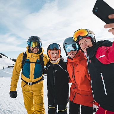 Eine Gruppe von vier Personen macht ein Selfie auf der Skipiste. Sie tragen bunte Skibekleidung und Helme, mit schneebedeckten Bergen im Hintergrund.